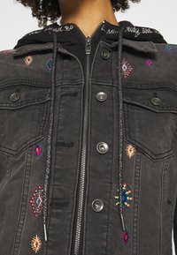 Svart denimjacka med färgglada broderidetaljer och en svart huva. Har tryckknappar och en frontdragkedja. Texturerad tyg.