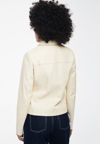 Crème fauxleren jas met een aansluitende silhouet, gekartelde kraag en strakke naaddetails, gecombineerd met donkerdenim broek.