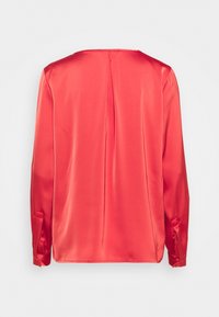 Vue arrière d'un chemisier en satin lisse de couleur rose corail à manches longues, avec un pli central et des poignets boutonnés sur un fond uni.