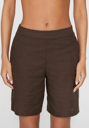 Mujer que lleva puestos unos pantalones cortos de lino marrón oscuro, de talle alto y largo hasta la rodilla, con una cintura ajustada, de pie con los brazos relajados a los lados.