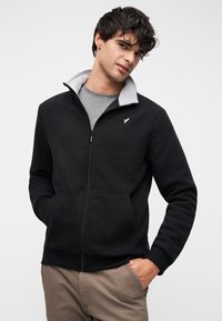 Μαύρη ζακέτα με φερμουάρ, γκρι εσωτερικό γιακά, υφή φλίς, πλαϊνές τσέπες και ένα μικρό λογότυπο στο αριστερό στήθος. Φορεμένη πάνω από γκρι πουκάμισο.