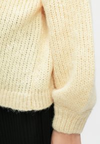 Manica di maglione lavorato a maglia color crema con polsino a coste, indossata sopra un tessuto nero a coste.