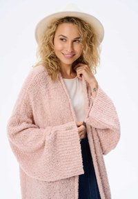 Różowy sweter z dzianiny z szerokimi rękawami, wykonany z teksturowanej włóczki. Posiada ściągacz na dole i widoczną metkę z przodu.