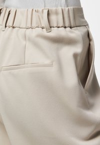 Gros plan sur un pantalon beige à taille élastique, passants de ceinture et une poche passepoilée à l’arrière, porté avec un haut blanc côtelé rentré.