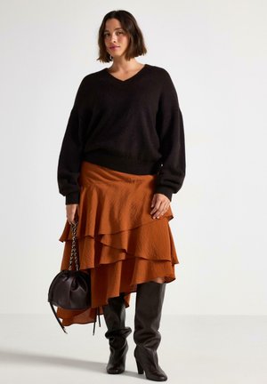 Maglione nero lavorato a maglia con scollatura a V, abbinato a una gonna arancione a balze. Il look è completato da stivali al ginocchio marroni con tacco e una borsa nera.