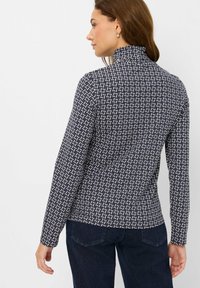 Maglietta a maniche lunghe blu navy con motivo geometrico bianco, colletto alto e design aderente, abbinata a jeans scuri.