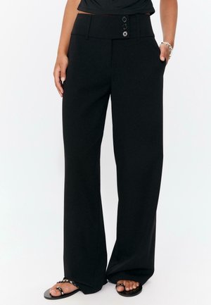 Pantalon noir à jambes larges, taille haute avec trois boutons, porté avec des sandales noires cloutées et un top noir, mains dans les poches.