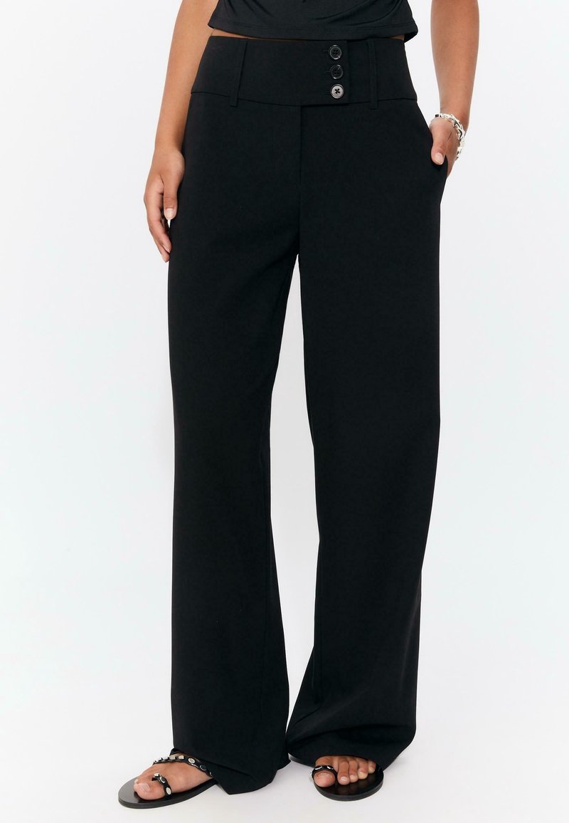 Pantalon noir à jambes larges, taille haute avec trois boutons, porté avec des sandales noires cloutées et un top noir, mains dans les poches.