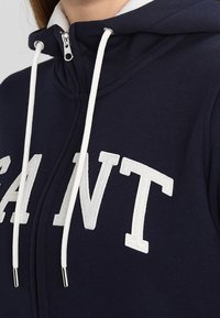 Felpa con zip di colore navy, caratterizzata da cordini bianchi e grandi lettere ricamate "SANT". Presenta un colletto alto e un tessuto morbido e texturizzato.