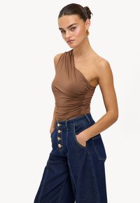 Haut marron à une épaule avec texture plissée, associé à un jean taille haute bleu foncé à jambes larges, mettant en avant des boutons dorés et des poches latérales.