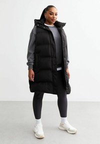 Gilet invernale nero con cappuccio, con un design trapuntato, bottoni a pressione e tasche laterali. Indossato sopra un felpa grigia e leggings.