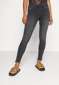 Jeans negros ajustados de mezclilla con una textura suave, combinados con sandalias planas marrones que presentan un diseño en capas y una suela negra.