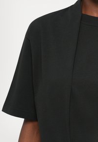 Detailný záber na osobu oblečenú v čiernej košeli s krátkymi rukávmi a otvorenej čiernej bunde na bielom pozadí.