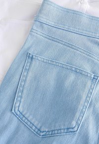 Ljusblå denim med en texturerad yta, som har en distinkt bakficka och kontrasterande vit tyg som syns ovanifrån.