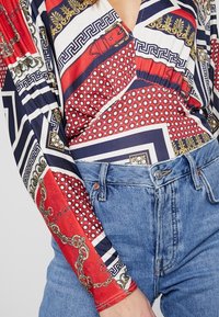 Langærmet bodysuit i multifarvet stof med geometriske mønstre, kæder og en v-halsdesign, parret med højtaljede denimjeans.