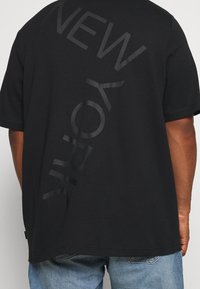 Czarna koszulka z dużym, tonalnym napisem "NEW YORK" z tyłu. Zaprojektowana w luźnym kroju i z krótkimi rękawami. Materiał to miękka bawełna.
