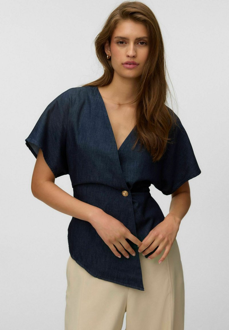 Femme aux longs cheveux bruns portant un haut cache-cœur bleu foncé à manches courtes avec un bouton et un pantalon beige, debout devant un fond clair uni.