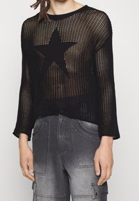 Schwarzer Strickpullover mit großem Sternmuster, offener Webstruktur, Rundhalsausschnitt und langen Ärmeln, kombiniert mit grauen Cargo-Hosen.