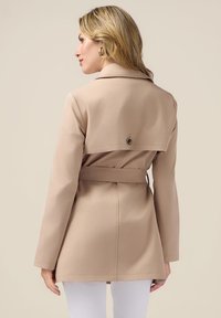 Beige trenchcoat met een ceintuur in de taille, lange mouwen en een gelaagd achterontwerp met een decoratieve knop en een gladde stoftextuur.