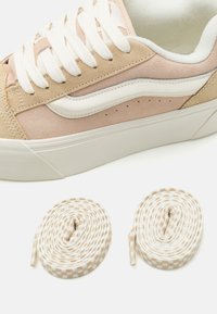 Light pink suede sneaker with white accents, featuring round laces and a textured sole. Two additional checkered laces are placed below.