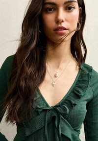 Mujer joven con cabello largo y oscuro, que lleva pendientes de aro dorados, collares dorados en capas y una blusa verde texturizada con cuello fruncido y lazo.