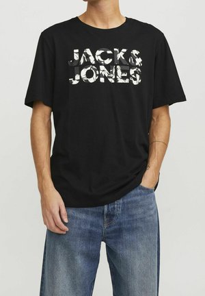 Mand iført sort T-shirt med korte ærmer med teksten "JACK & JONES" i hvid og blå jeans mod en lyst grå baggrund.