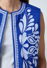 Blå broderet vest med indviklede hvide blomsterdekorationer, dekorative syninger langs kanterne og ærmeløs design.