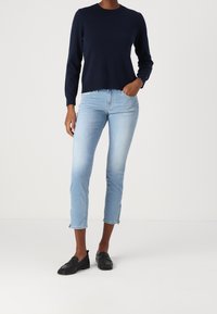 Mujer que lleva un suéter de manga larga azul marino, vaqueros cortos de color azul claro con cremalleras en los tobillos y zapatos de loafers negros sin cordones, de pie con una mano en el bolsillo.