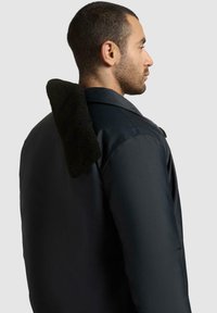 Sort jakke med en strukturert, mørkegrønn fleecekrage. Glatt stoff og skreddersydd design med synlige sømmer. Sideprofil.