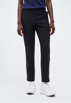 Persoană purtând pantaloni negri, strâmți, adidași albi și un top albastru închis, stând pe un fundal simplu, deschis la culoare.