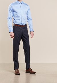 Man med ljusblå skjorta, mörkblå kostymbyxor, brun läderbälte och bruna finskor som står inomhus.