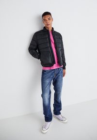 Giacca a vento nera con colletto alto, indossata sopra una camicia rosa. Jeans blu e sneakers viola chiaro completano l'outfit. Texture liscia.