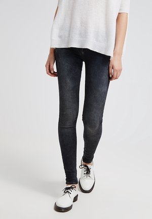 Persoon die versleten zwarte skinny jeans, een wit gebreide trui en witte veterschoenen draagt en loopt op een effen vloer.
