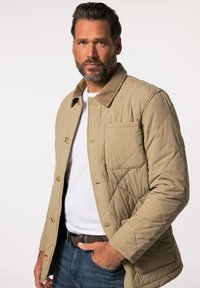 Giubbotto imbottito beige con colletto in corduroy, quattro bottoni frontali e tasca sul petto. Indossato sopra una camicia bianca e jeans blu.