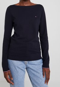 Kvinna som bär en marinblå långärmad tröja med liten logotyp och ljusblå jeans, stående mot en enfärgad bakgrund.