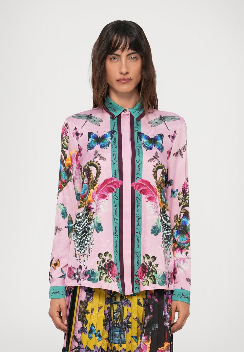 Vrouw in een roze blouse met kleurrijke vlinders, libellen, bloemen- en veermotieven, gecombineerd met een geplooide, multikleurige rok.