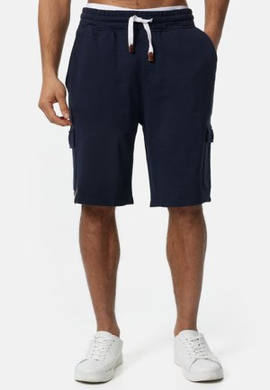 Mann trägt marineblaue Cargo-Shorts mit weißem Kordelzug und weiße Turnschuhe, steht vor weißem Hintergrund.