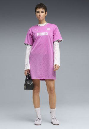 Młoda osoba ubrana w różową sportową sukienkę Puma na białą koszulę z długim rękawem, białe skarpety, sportowe buty oraz trzymająca czarną torebkę.