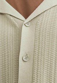 Ljus beige stickad kofta med en texturerad mönster, bred krage och tre vita knappar fram. Mjuk tyg med ribbade detaljer.