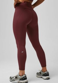 Bordeauxfarbene, hochtaillierte Leggings mit kontrastierenden Nähten, figurbetonter Passform und kleinem Logo an der unteren Wade, kombiniert mit schwarz-weißen Laufschuhen.