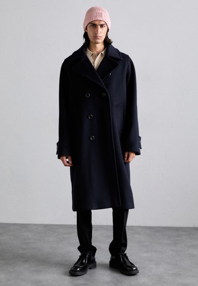 Manteau en laine bleu marine à double boutonnage avec de grands boutons, poches latérales et longue longueur, associé à un bonnet en maille côtelée rose clair et des chaussures noires.