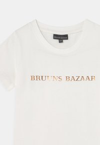T-shirt branca de manga curta com um decote redondo, apresentando uma impressão bronze brilhante que diz "BRUUINS BAZAAR" no peito.