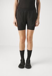 Pantalones cortos deportivos negros con cintura elástica, superpuestos sobre pantalones cortos ajustados negros. Combinados con calcetines negros y zapatillas de correr.