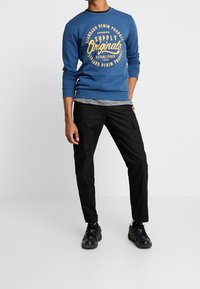 Blå sweatshirt med gul grafisk text, grårandig skjorta under, svarta cargobyxor och svarta sportskor. Standard passform silhuett.