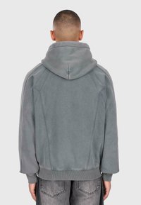 Personne avec les cheveux courts portant un sweat à capuche gris et un jean noir délavé, de dos contre un fond clair et uni.