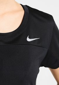 Camisa atlética negra con cuello redondo, que presenta un panel de malla texturizada en los hombros y un logo de Nike en blanco en el lado izquierdo.