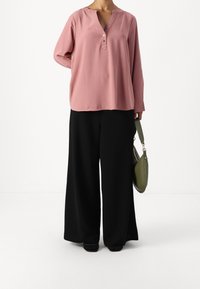 Prachově růžový top s výstřihem do V a tříčtvrtečními rukávy, ozdobený knoflíkem, spárovaný s černými širokými kalhotami a zelenou kabelkou přes rameno.