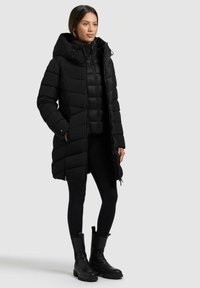 Svart dunjakke med lang design, trekantete quilted mønster, justerbar hette og glidelåslommer. Båret over svarte leggings og klumpete støvler.