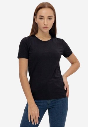 Donna con capelli castani lisci che indossa una semplice maglietta nera a maniche corte e jeans blu, in piedi con una mano sul fianco.