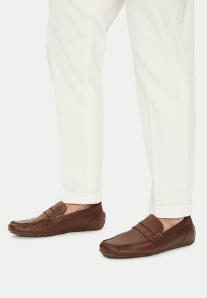 Mocasines de piel marrón con parte superior texturizada, detalles de costura y una banda frontal elevada, combinados con pantalones blancos arremangados.